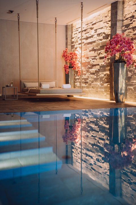 Indoor-Pool im Wellnessbereich mit schwebender Liege, Steinwand und rosa Orchideen.