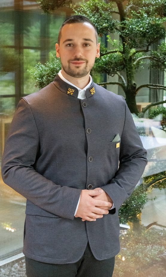 Hotelmitarbeiter in grauer Uniform steht vor einer Glasfront mit dekorativem Baum im Innenraum.