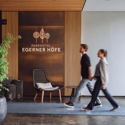 Ein Paar geht durch den Eingangsbereich des Parkhotel Egerner Höfe mit Holzlamellen, Pflanze und Sessel.