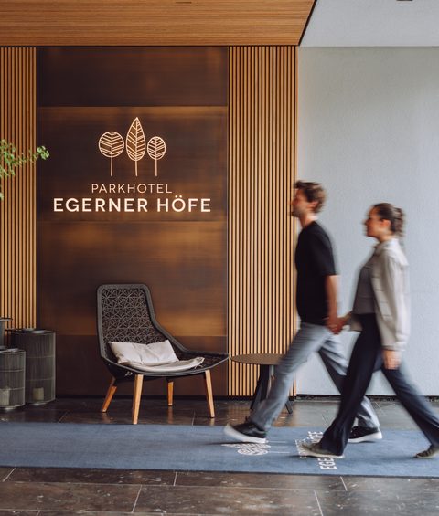 Ein Paar geht durch den Eingangsbereich des Parkhotel Egerner Höfe mit Holzlamellen, Pflanze und Sessel.