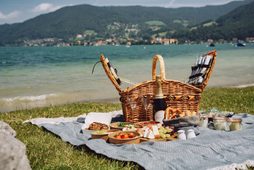 Picnic basket with food and sparkling wine on a blanket by the lakeshore, with water and mountains behind.