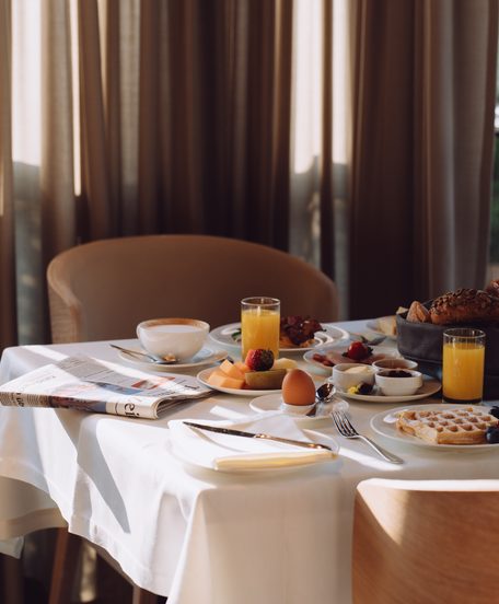 Gedeckter Frühstückstisch mit Kaffee, Orangensaft, Gebäck, Obst und Waffeln vor hohen Vorhängen.