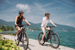 Zwei Personen fahren mit Fahrrädern auf einer Uferpromenade an einem See mit Bergen im Hintergrund.