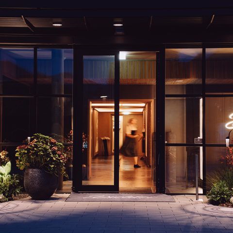 Illuminated hotel entrance with glass facade, plants, and view into the lobby at night.