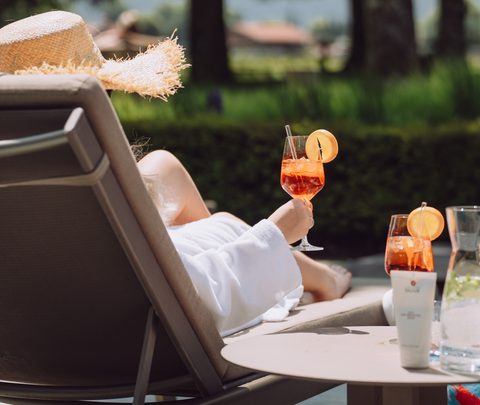 Person im Bademantel auf einer Liege im Außenbereich mit zwei Aperol Spritz auf einem Tisch.