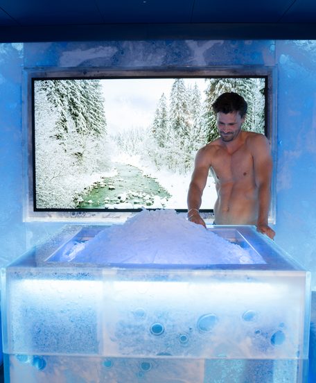 Zwei Gäste in Badekleidung in einem blau beleuchteten Eisraum mit Schnee auf einem Eistisch und Winterblick.