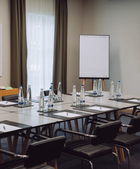 Seminarraum mit U-förmig gestellten Tischen, Stühlen, Wasserflaschen, Schreibblöcken, Flipchart und Fernseher.
