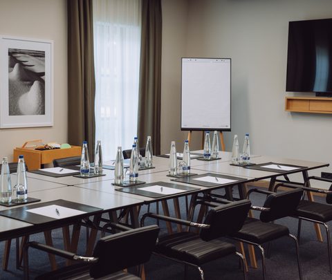 Seminarraum mit U-förmig gestellten Tischen, Stühlen, Wasserflaschen, Schreibblöcken, Flipchart und Fernseher.