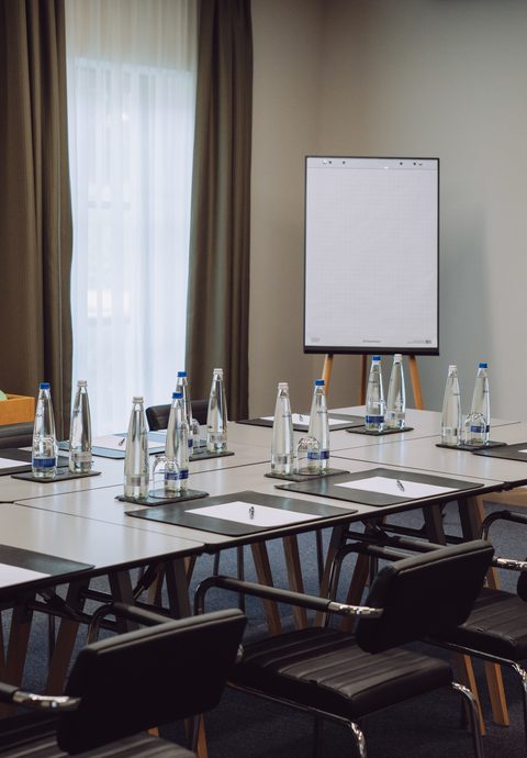 Seminarraum mit U-förmig gestellten Tischen, Stühlen, Wasserflaschen, Schreibblöcken, Flipchart und Fernseher.