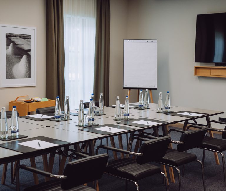 Seminarraum mit U-förmig gestellten Tischen, Stühlen, Wasserflaschen, Schreibblöcken, Flipchart und Fernseher.
