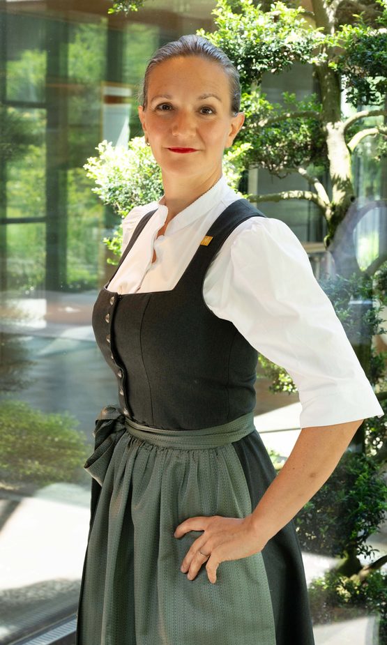 Hotelmitarbeiterin in traditioneller Uniform steht vor Glasfront und dekorativen Pflanzen im Innenbereich.