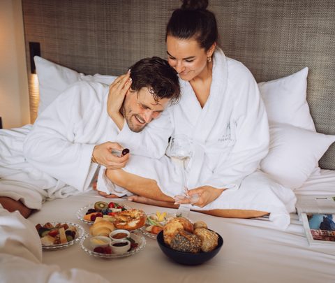 A couple in bathrobes sits on a hotel bed enjoying breakfast with pastries, fruit and drinks.