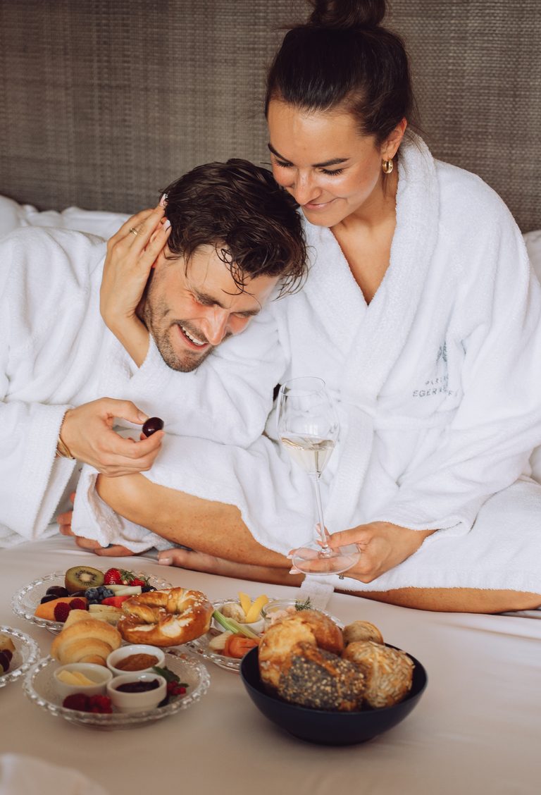 Ein Paar im Bademantel sitzt auf einem Hotelbett und genießt Frühstück mit Gebäck, Obst und Getränken.