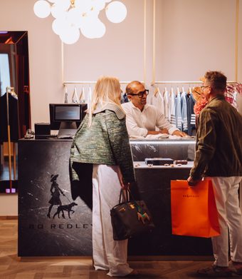 Zwei Gäste stehen mit einem Verkäufer an der Theke in einer Boutique mit Kleidung und Spiegel.