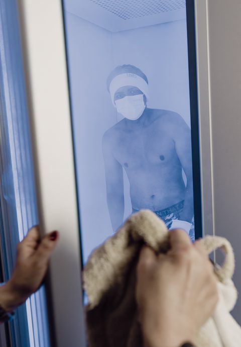 A woman opens a cryotherapy chamber where a man stands wearing a mask and headband.