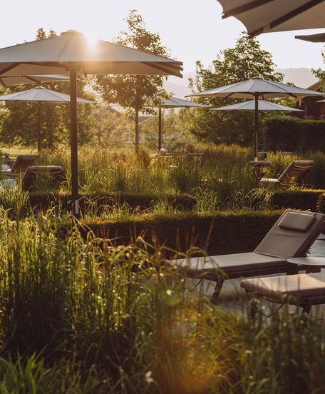 Sonnenliegen und Sonnenschirme auf einer Wellness-Terrasse in einem Garten bei tief stehender Sonne.