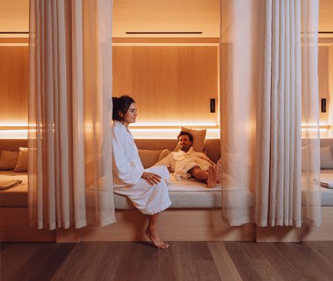 Ein Paar in Bademänteln entspannt im Ruheraum eines Wellnessbereichs zwischen Vorhängen auf Liegen.