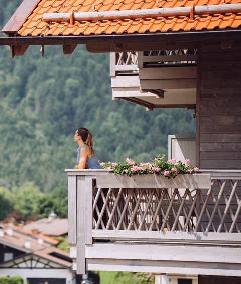 Eine Frau steht auf einem Hotelbalkon mit Blick auf Dorf und bewaldeten Hang.