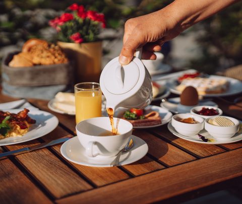 Eine Hand gießt Kaffee aus einer Kanne in eine Tasse auf einem gedeckten Frühstückstisch auf der Terrasse.