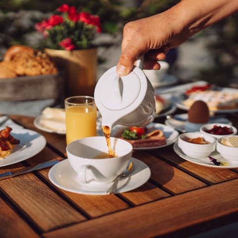 A hand pours coffee from a pot into a cup on a set breakfast table on the terrace.
