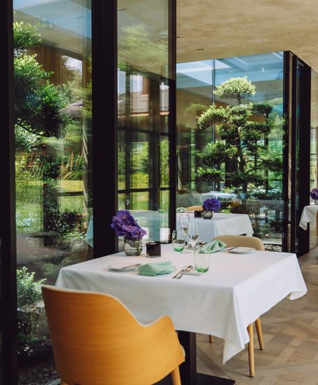 Restaurant mit weiß gedeckten Tischen an einer großen Fensterfront mit Blick in den Garten.