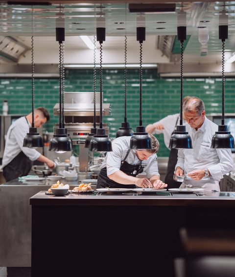 Mehrere Köche richten in einer offenen Restaurantküche unter Wärmelampen Speisen an.