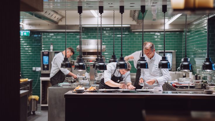 Mehrere Köche richten in einer offenen Restaurantküche unter Wärmelampen Speisen an.