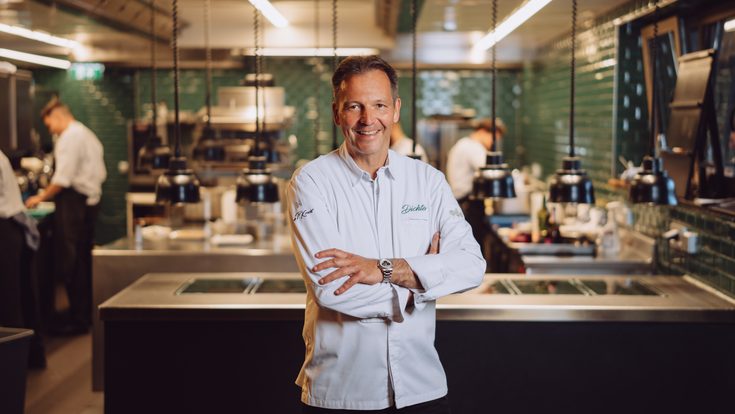 Koch in weißer Jacke steht in einer offenen Restaurantküche vor Arbeitsflächen und Küchenteam im Hintergrund.