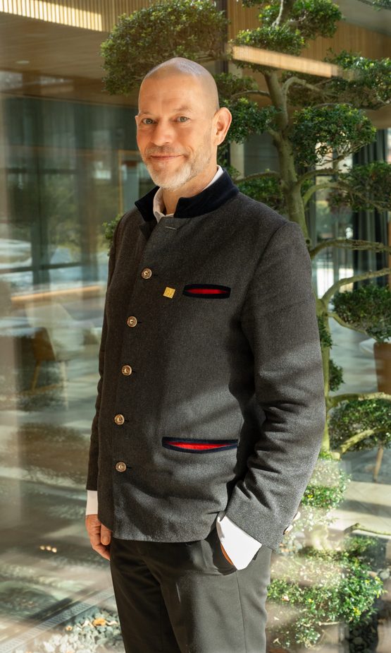 Hotelmitarbeiter in dunkler Uniform steht in einer hellen Lobby vor einer geformten Pflanze.