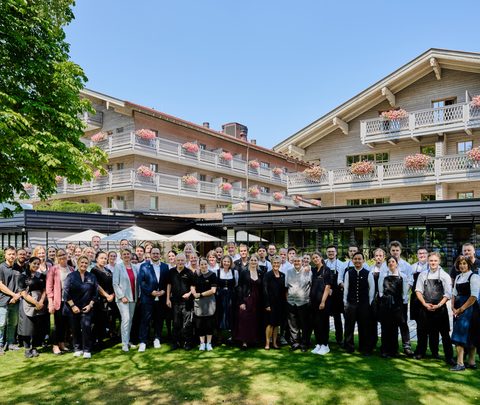 Gruppenfoto des Hotelteams im Garten vor einem Hotelgebäude mit Balkonen und Blumen.