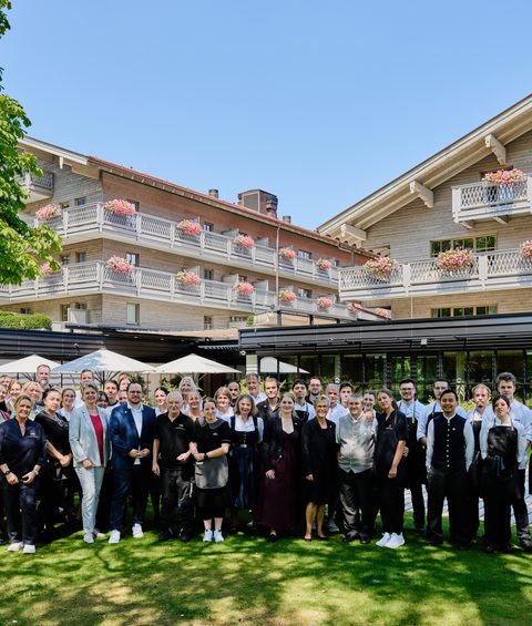 Gruppenfoto des Hotelteams im Garten vor einem Hotelgebäude mit Balkonen und Blumen.