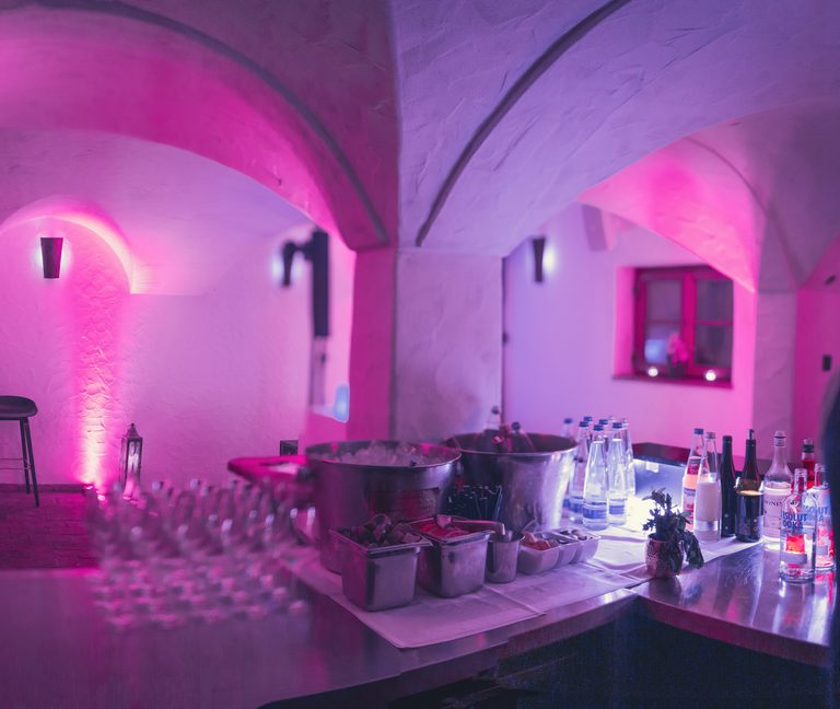 Gewölbekeller mit Barbereich, Getränkeflaschen und pinker Beleuchtung an Tresen und Sitzplatz.
