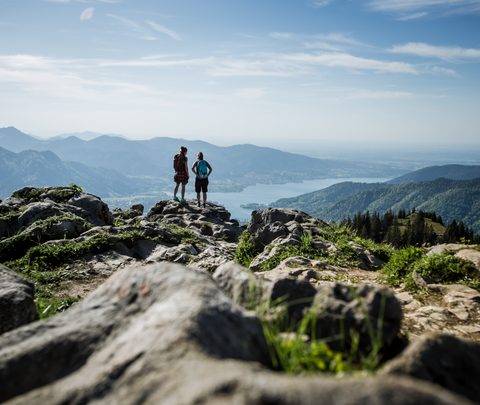Two hikers stand on a rocky mountain ridge overlooking mountains, valley and lake.