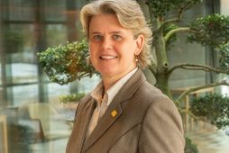 Hotel staff member in business attire standing indoors by a glass wall and decorative tree.
