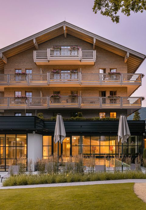 Außenansicht eines Hotels mit Balkonen, Terrasse und Garten in der Abenddämmerung.