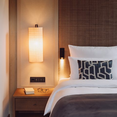 Detail eines Hotelzimmers mit Bett, Nachttisch, Buch, Brille und warm beleuchteter Wandlampe.