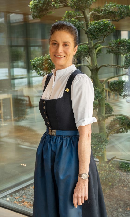 Hotelmitarbeiterin in Tracht steht lächelnd vor einer Glasfront neben einem dekorativen Baum im Innenbereich.