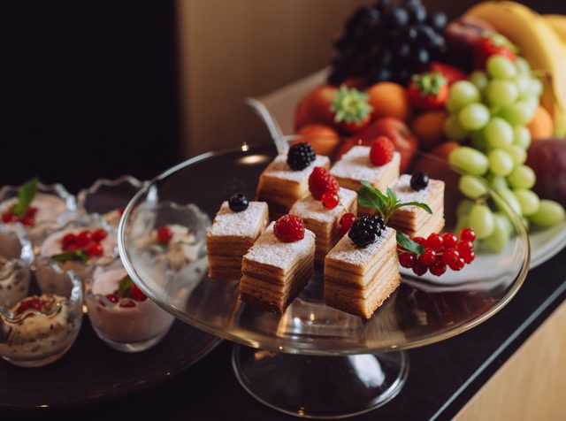 Kuchenstücke und Dessertgläser auf einem Buffet neben einer Obstplatte mit Trauben und Bananen.