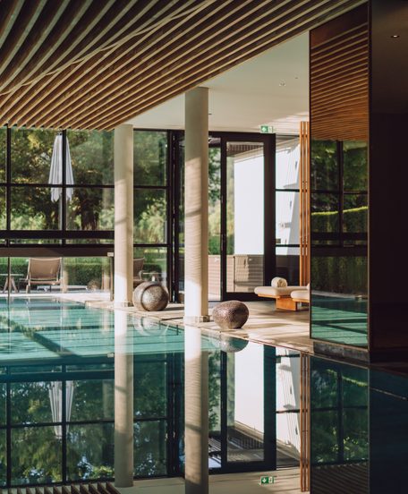 Innenpool im Wellnessbereich mit Ruheliegen, großer Fensterfront und Holzdecke.