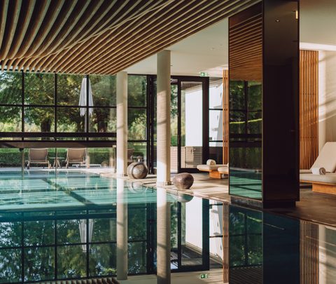 Innenpool im Wellnessbereich mit Ruheliegen, großer Fensterfront und Holzdecke.