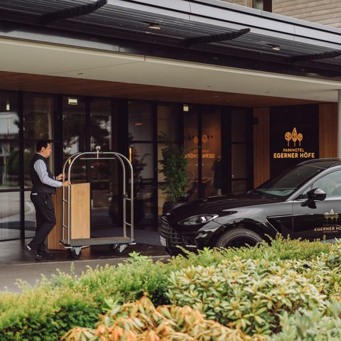Hotel staff member with luggage cart at a covered hotel entrance beside a black car.