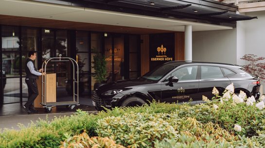 Hotel staff member with luggage cart at a covered hotel entrance beside a black car.