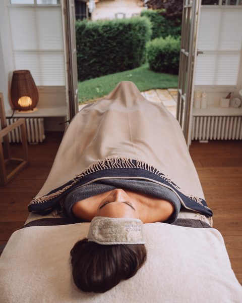 Frau auf einer Wellnessliege in einem Behandlungsraum mit offenen Türen und Blick in den Garten.
